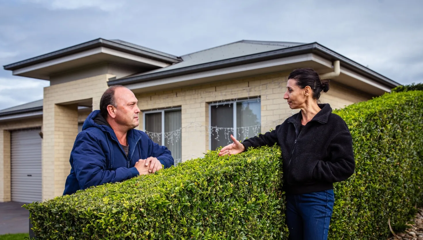 Two neighbours talking over a fence with a house in the background, representing community awareness.