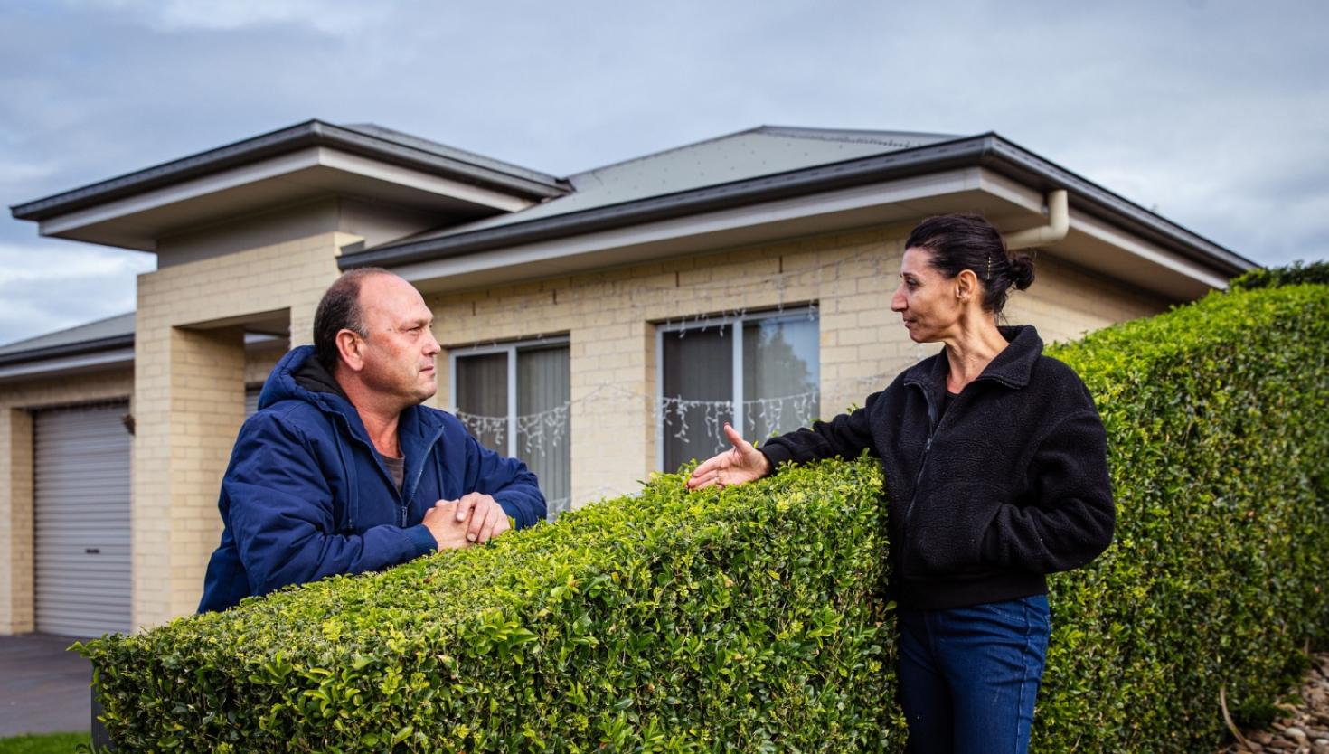 Two neighbours talking over a fence with a house in the background, representing community awareness.
