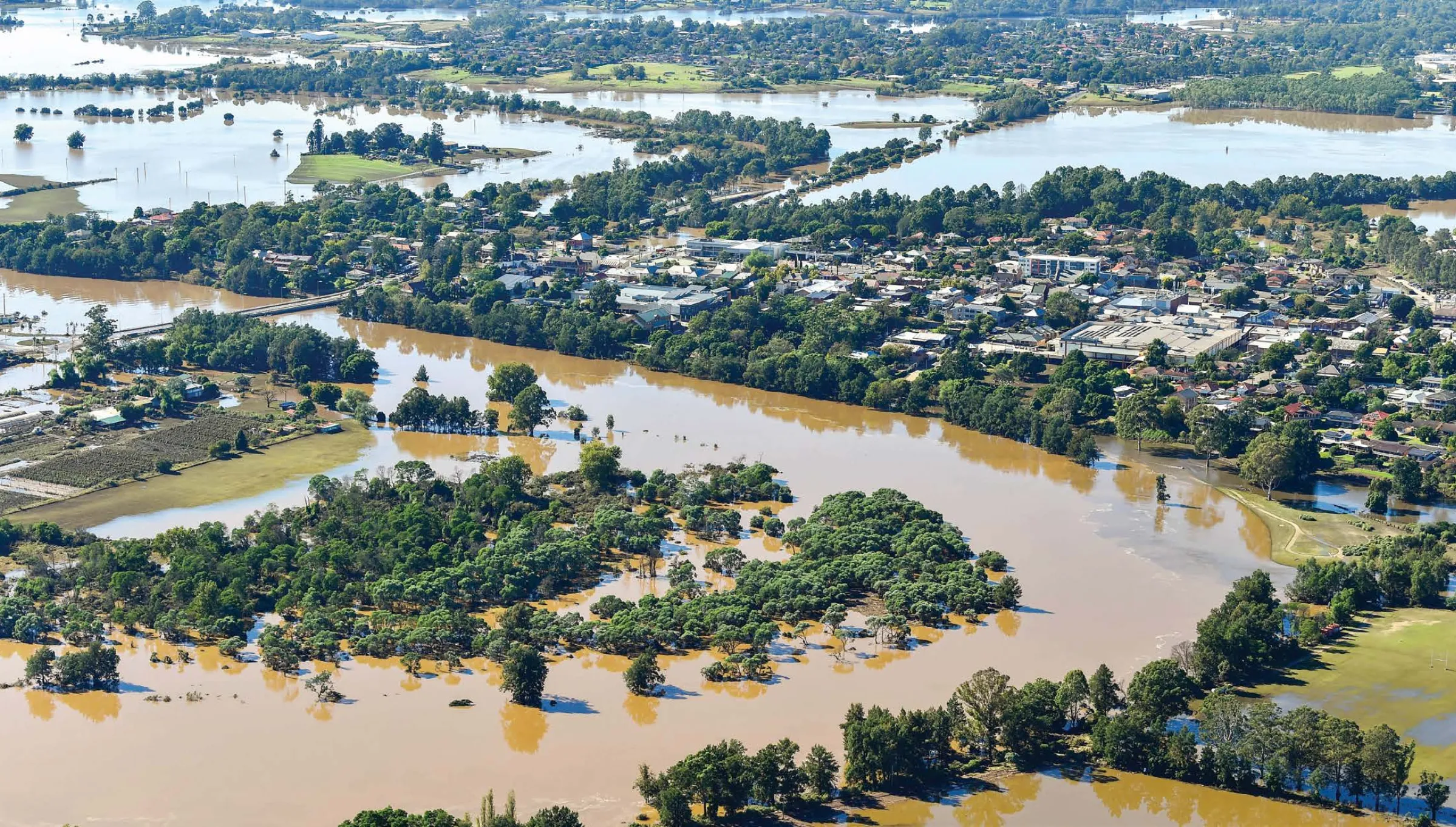Flooded landscape in the Hawkesbury-Nepean area of NSW, with fields and trees partially submerged under murky floodwater.