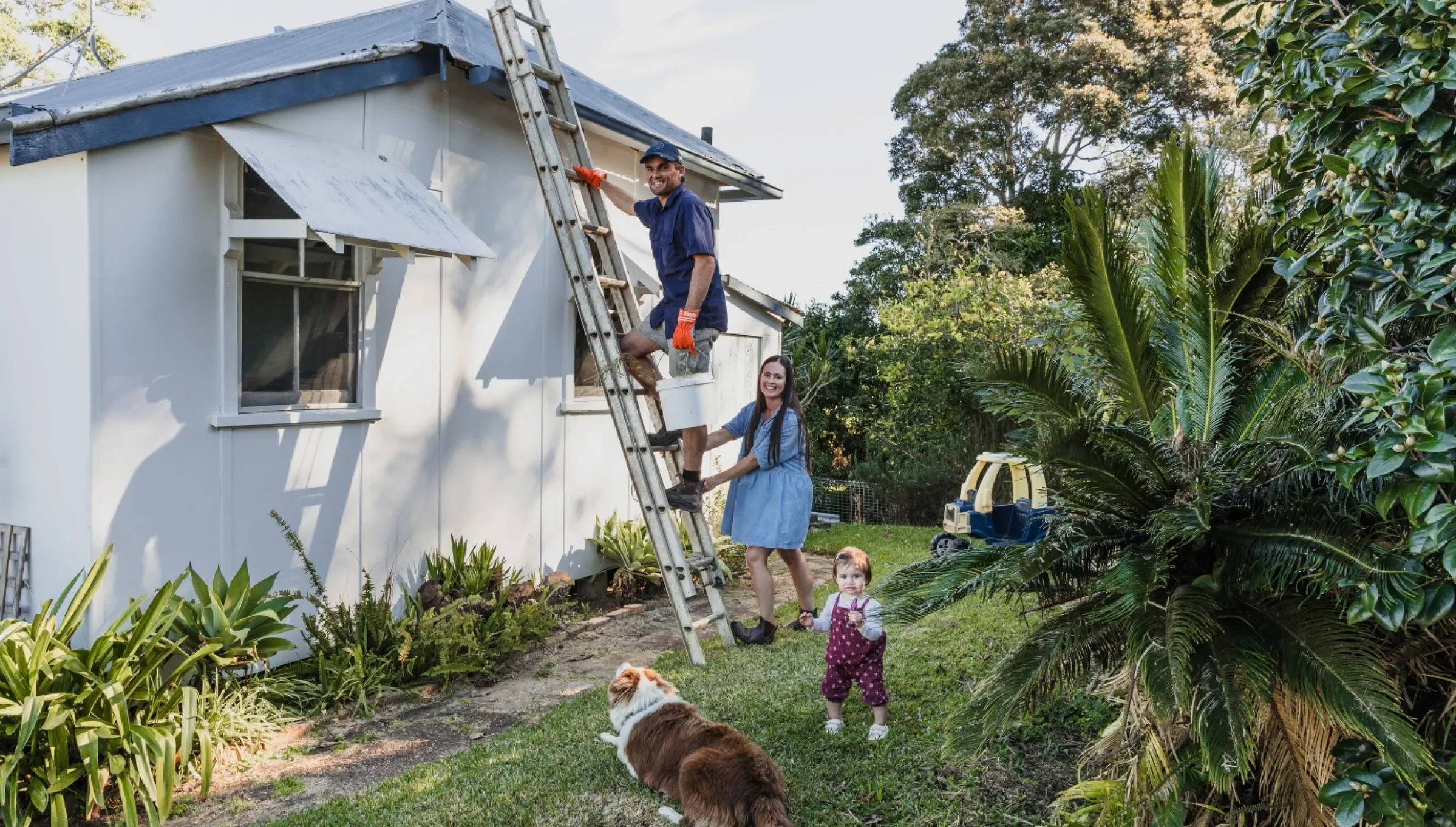 Man climbs a ladder to work on the house roof, while a woman holds the ladder steady. A family prepares for storm season together, with a baby and a dog nearby, watching attentively.