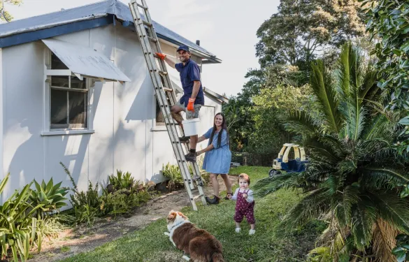 Man climbs a ladder to work on the house roof, while a woman holds the ladder steady. A family prepares for storm season together, with a baby and a dog nearby, watching attentively.