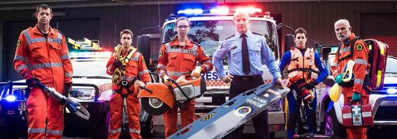 Six NSW SES members in orange uniforms holding rescue tools in front of a NSW SES truck and vehicles..