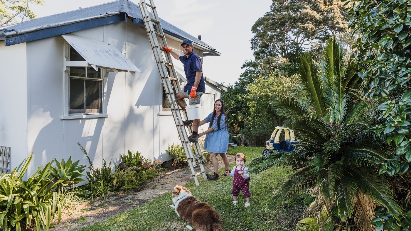 Man climbs a ladder to work on the house roof, while a woman holds the ladder steady. A family prepares for storm season together, with a baby and a dog nearby, watching attentively.